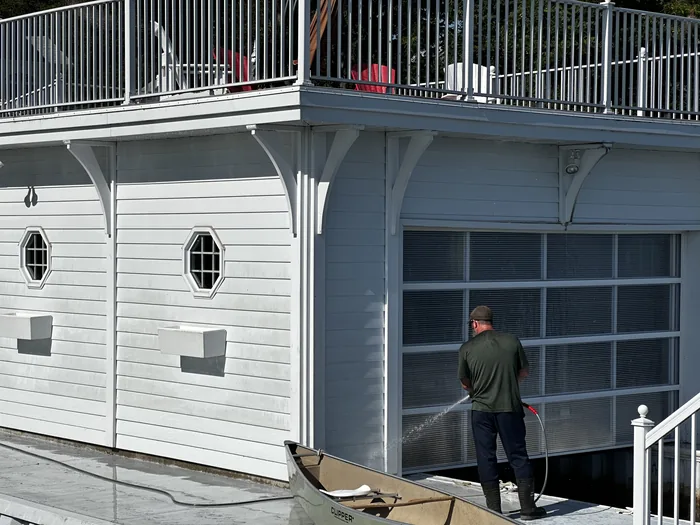 A Nord tech soft-washing the clapboard wall of a two-storey boathouse, the low-pressure spray fanning across the siding beside the slip