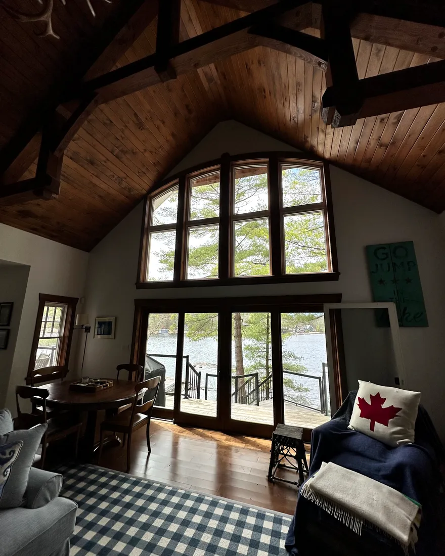 The great room of a Simcoe County cottage — timber ceiling, tall arched window, and the lake and dock clearly visible through freshly cleaned glass