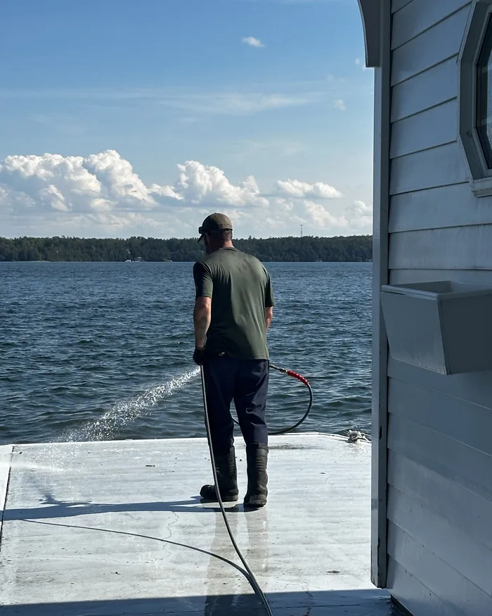 A Nord tech rinsing a lakeside dock, the spray fanning across wet boards with open water and summer cloud behind