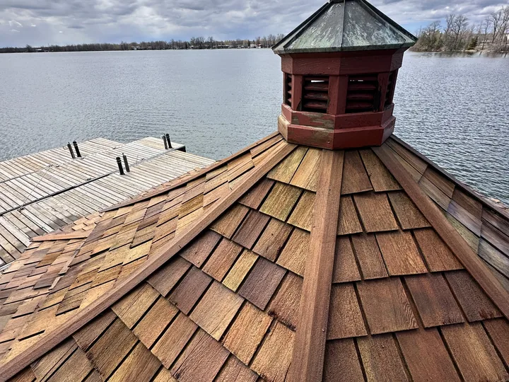 The cedar-shingled roof of a lakeside boathouse restored to warm brown, its copper-topped cupola and open water behind