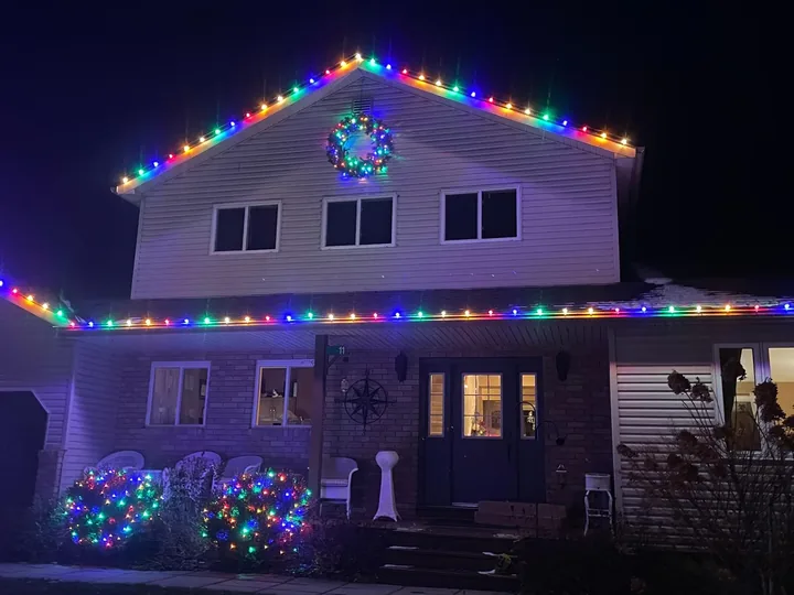 A home outlined in multi-colour C9 roofline bulbs with a lit wreath in the gable and colour-lit shrubs below