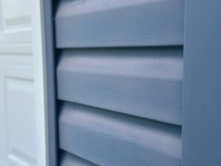 A close-up of blue vinyl siding whose surface has gone flat and chalky pale under years of sun