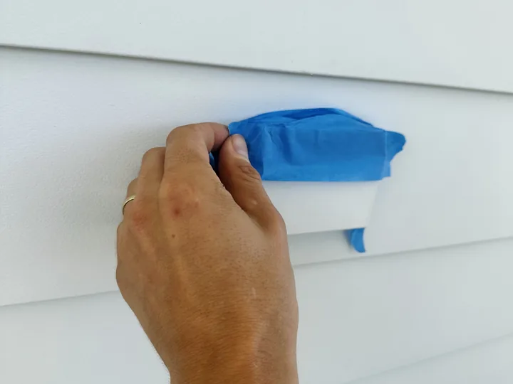 A hand pressing painter's tape over an exterior electrical receptacle on white siding, sealing it before a soft wash begins