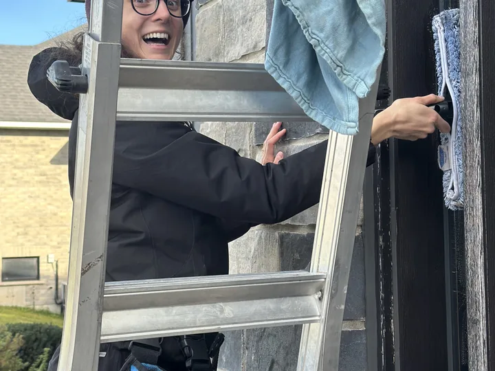 A Nord window cleaner on a ladder at a stone-faced home, scrubber in hand and a tool bag hooked to the rungs