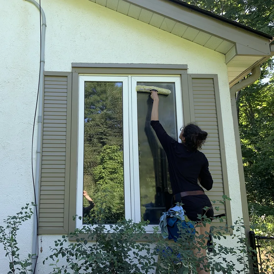 Liz of Nord, up a ladder at a cream stucco home, washing the upper pane of a tall window with a scrubber