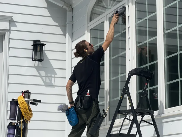 A Nord window cleaner squeegeeing tall arched windows from a step ladder, water-fed pole cart and buckets set up on the patio beside him